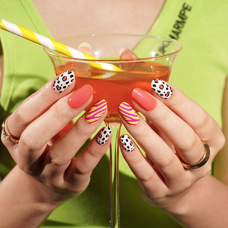Hands holding cocktail glass showcasing retro eye pattern nail wraps with coral pink nails