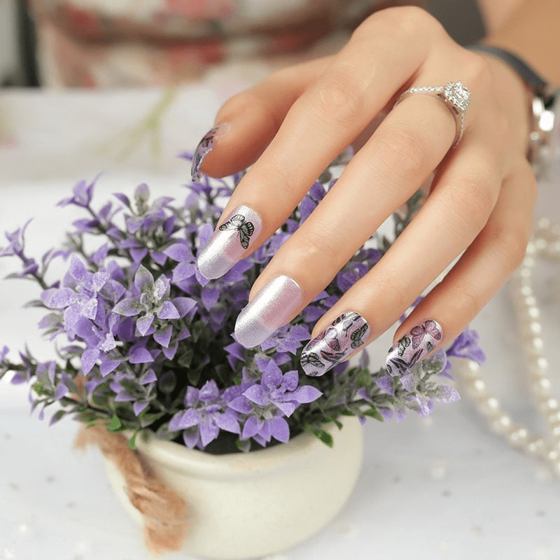 Hand with purple butterfly nail wraps reaching for lilac flowers showing at-home manicure