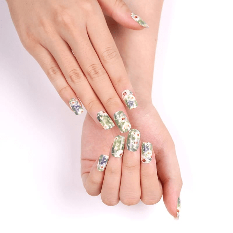 Two hands showcasing white floral nail wraps with green daisy botanical patterns