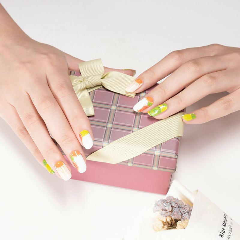 Hands applying citrus nail wraps with yellow and orange fruit patterns near gift box