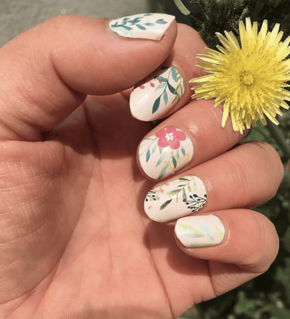 Hand with yellow flower showing cream nail wraps featuring pink and green botanical floral motifs