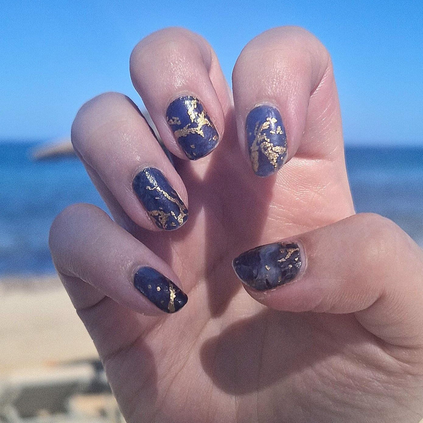 Royal blue marble nail wraps with gold veining on hand against beach background