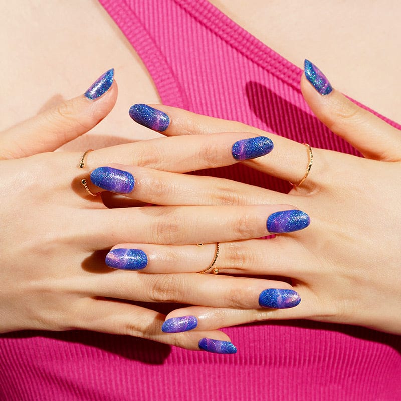 Hands with magnetic blue purple nail wraps displaying holographic chrome effect against pink background