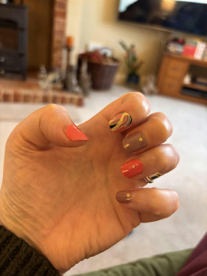 Hand displaying coral and brown nail wraps with gold foil feather pattern at-home manicure