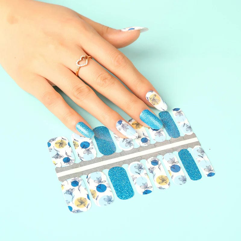 Hand holding botanical non-UV nail wraps sheet featuring blue glitter and illustrated leaf nail art designs