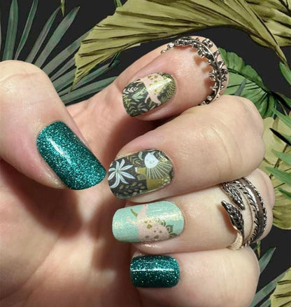 Green glitter and dinosaur patterned nail wraps on hand with botanical leaf designs