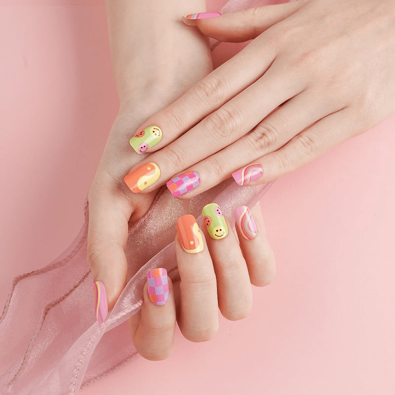 Hand showing 90s-inspired nail wraps with smiley faces, checkered print, and rainbow swirl designs