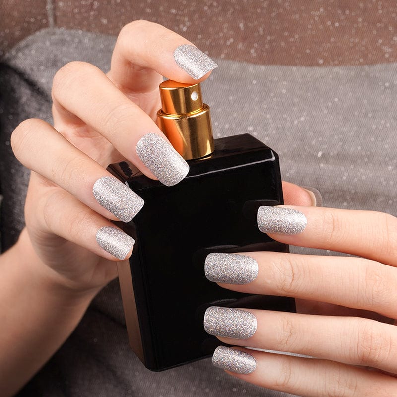 Hands holding perfume bottle showcasing silver shimmer nail wraps for glamorous manicure