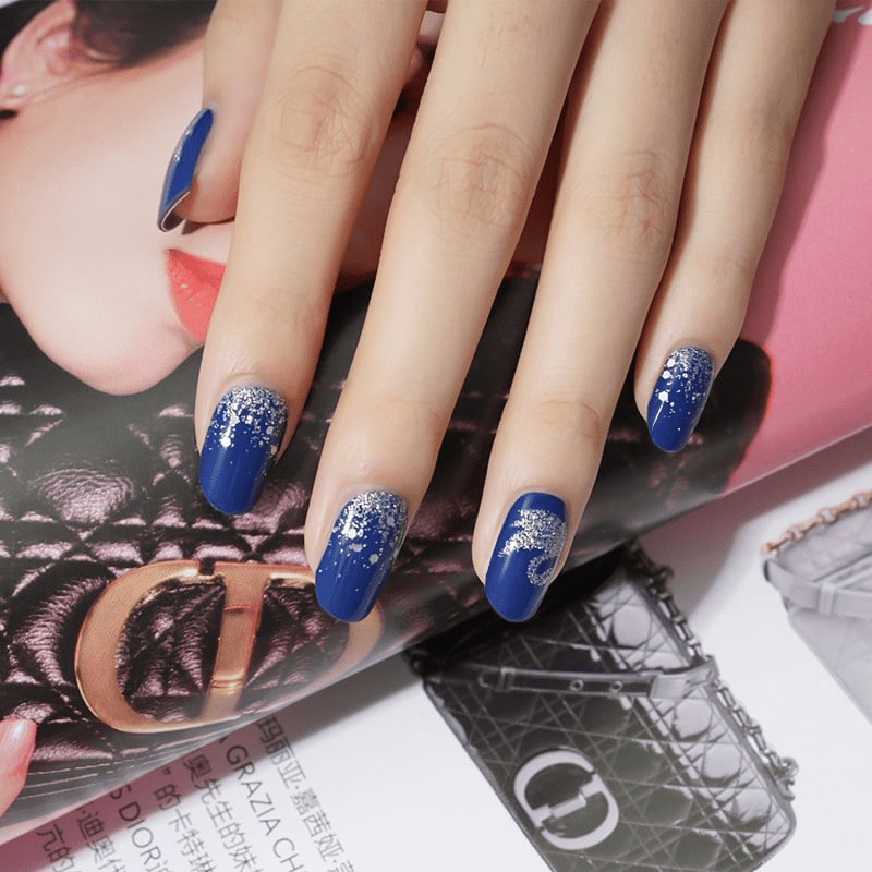 Navy blue nail wraps with silver swan patterns displayed on hand over magazine