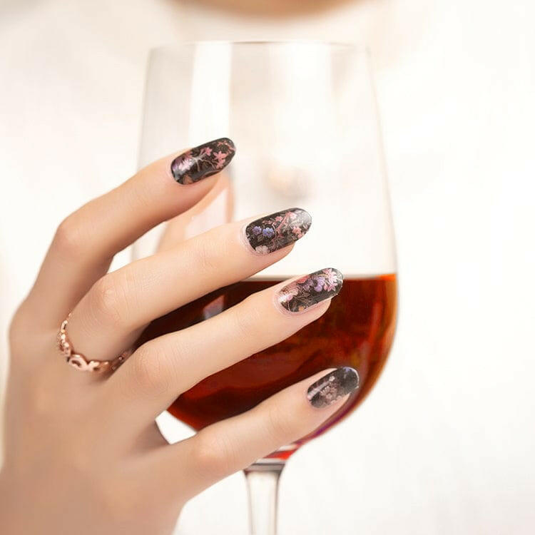 Black floral gel nail wraps on manicured hand holding wine glass with dark botanical design
