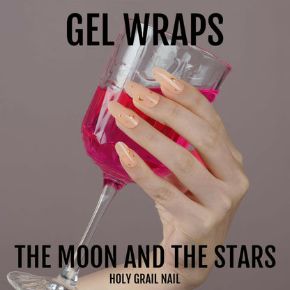 Hand holding pink wine glass wearing peachy gel nail wraps with gold celestial designs